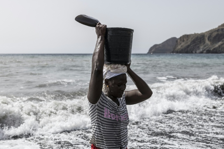 Vania Tavares porte en équilibre sur sa tête un seau rempli de sable noir, sur une plage dégradée par l'exploitation commerciale, près de Ribeira da Barca, le 20 mai 2026 au Cap-Vert