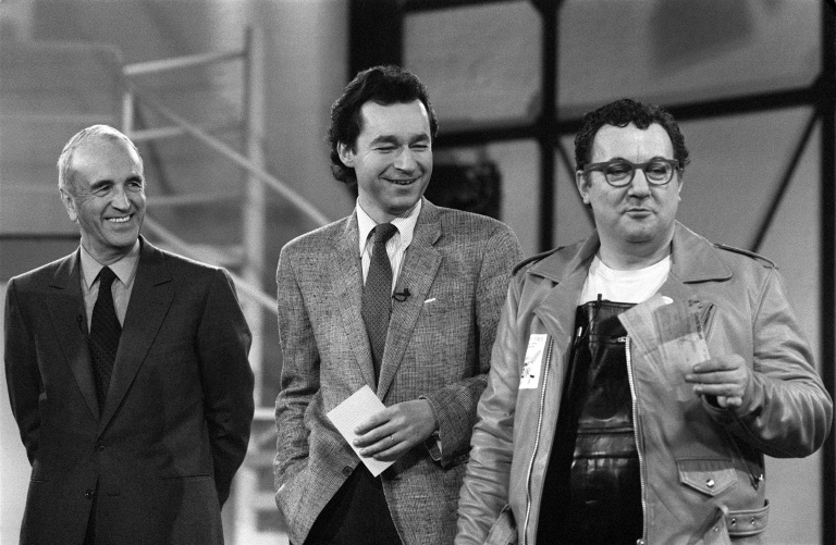 Coluche, à droite, montre le 29 janvier 1986 à Paris le chèque de 205.000 francs collectés au profit des Restos du Coeur en compagnie d'André Rousselet, fondateur de la chaîne cryptée Canal, à gauche, et de Michel Denisot, futur directeur des Sport de Canal, au centre