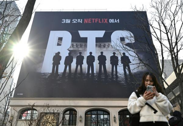 Une femme consulte son smartphone devant un panneau d'affichage annonçant le concert de retour du groupe de K-pop BTS, le 17 mars 2026 à Séoul