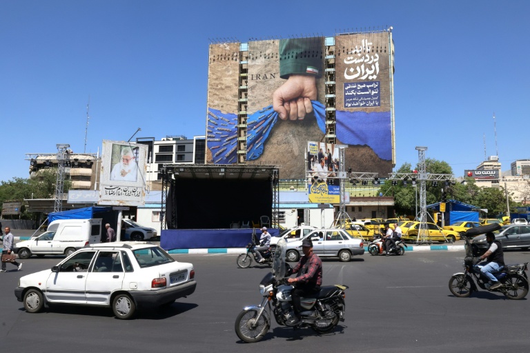 Des véhicules devant un panneau d'affichage sur la façade d'un bâtiment représentant le détroit d'Ormuz avec une légende en persan disant 