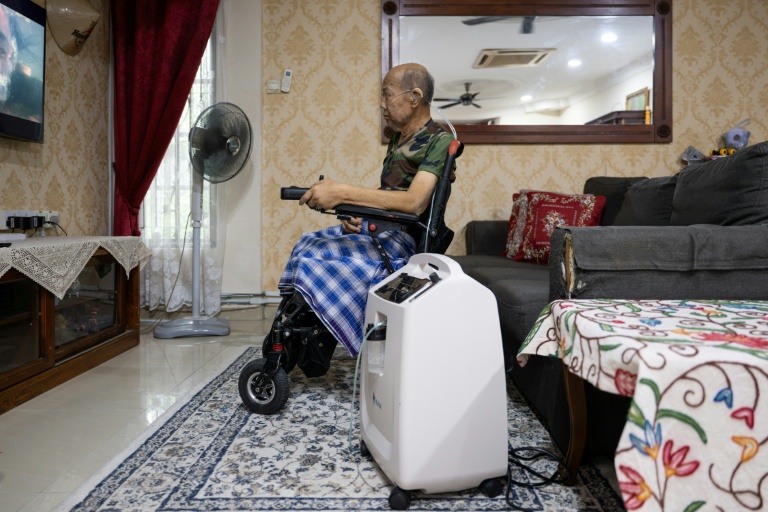 Md Noor Bin Bujang regarde la télévision près d'un concentrateur d’oxygène à son domicile de Rawang, dans l’État de Selangor, le 16 octobre 2026 en Malaisie