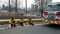 Trois membres du service d'incendie de Spokane en pause sur le bord du trottoir devant des maisons d'habitation incendiées, dans le quartier de Balboa à Spokane (Washington), le 2 août 2026